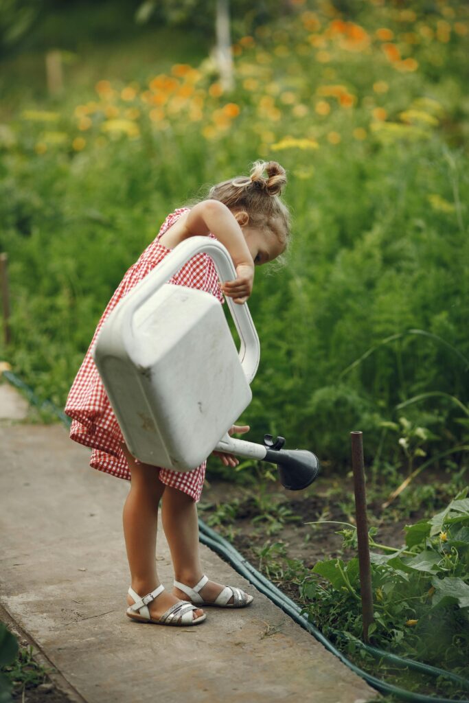 little girl in gingham dress and white sandals using a watering can to water plants